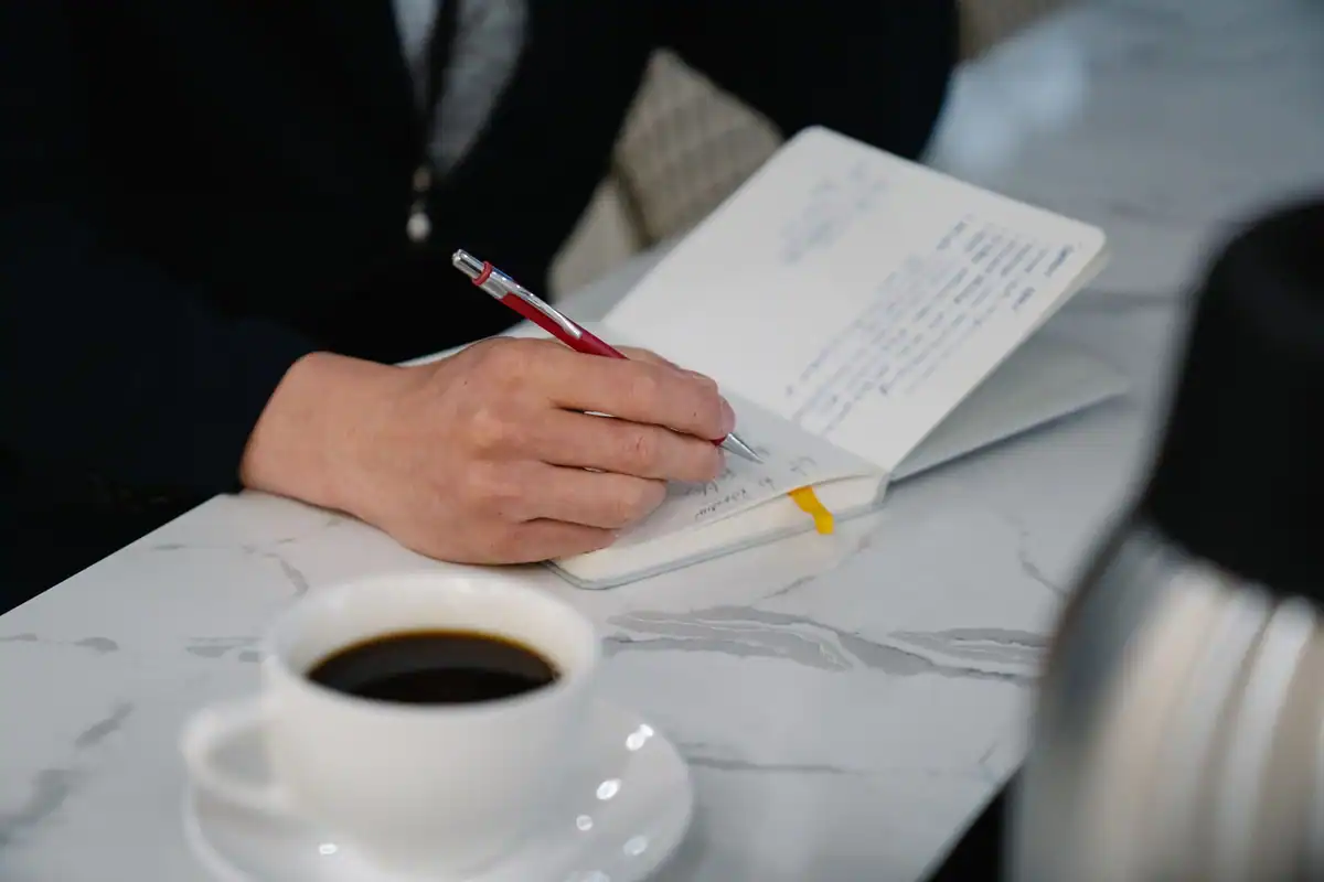 Mână care scrie într-un jurnal de lucru, lângă o cafea neagră