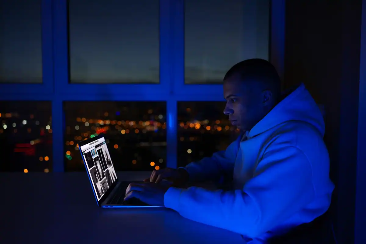 Bărbat care lucrează seara pe laptop, lângă o fereastră cu luminile orașului
