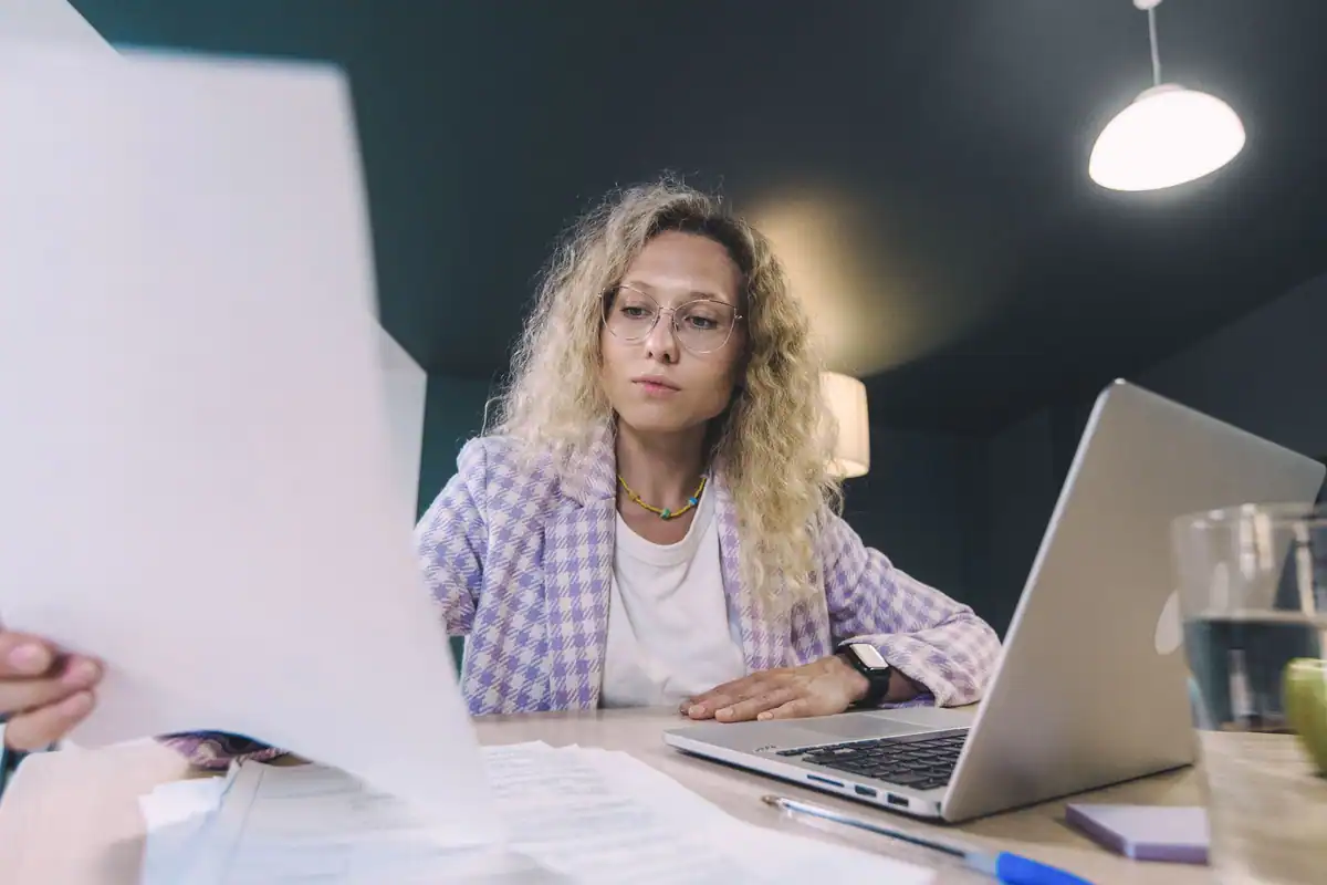Femeie care analizează documente în fața laptopului, într-un birou cu pereți închiși la culoare
