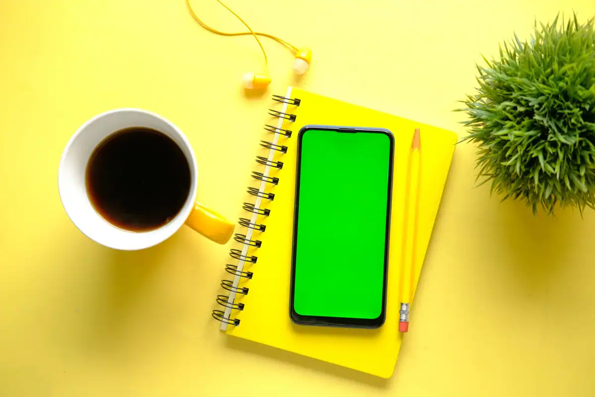 Birou galben minimalist cu caiet, telefon, cafea și o plantă