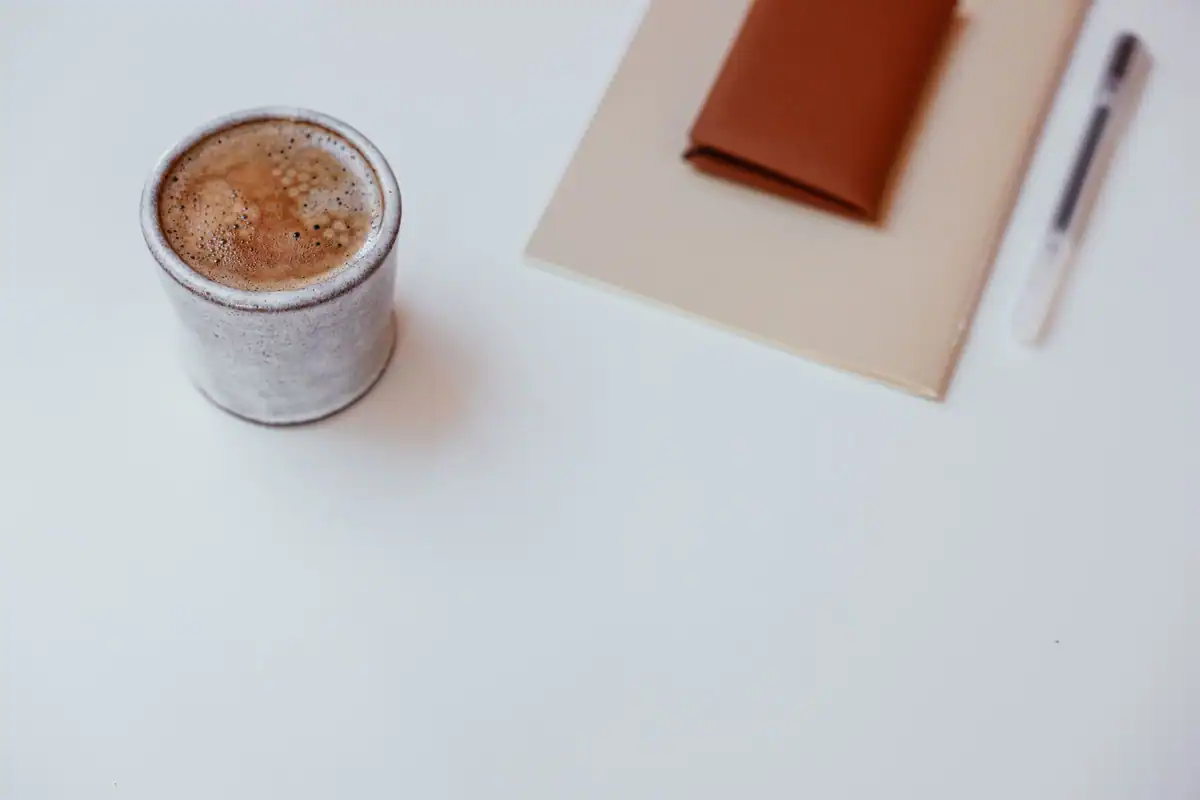 Ceașcă de cafea, caiet și pix pe o masă albă, într-un decor minimalist
