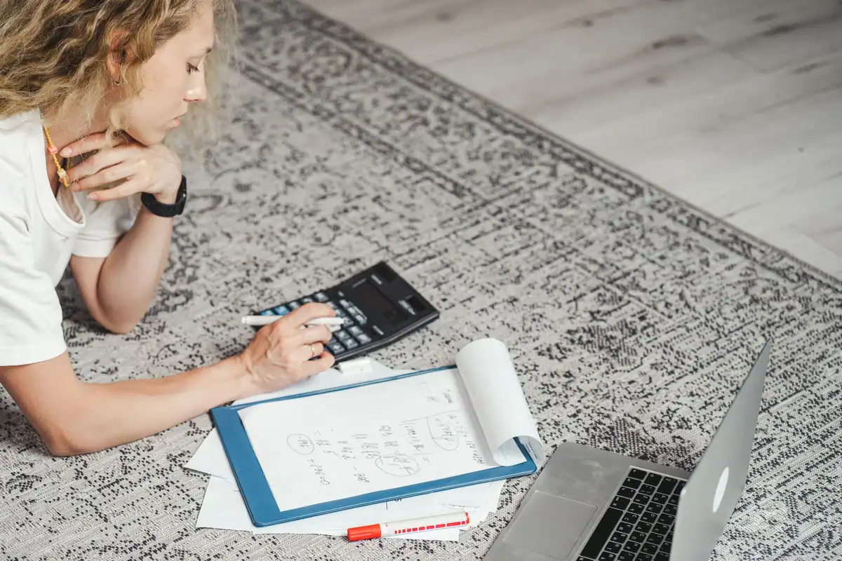 Femeie întinsă pe covor care calculează un tarif pe calculator, cu laptopul alături